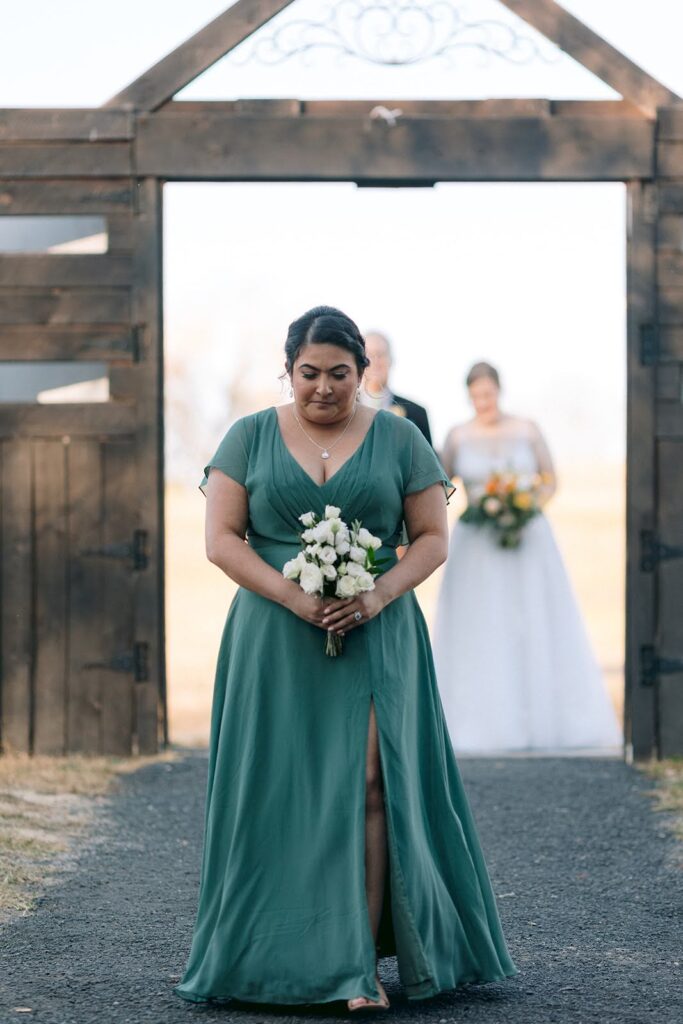 A bridesmaid walks down the aisle at Arbor Haven in Fredericksburg,VA. She wears a soft blue-green dress and carries a white and green small bouquet. Flowers by One Last Avocado Floral Design.