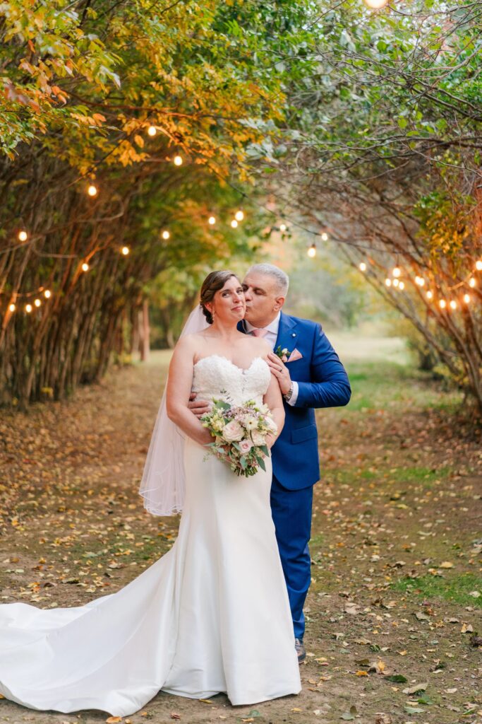 A bride and groom stand in a tree tunnel with fairy lights. The trees are beginning to turn orange.
