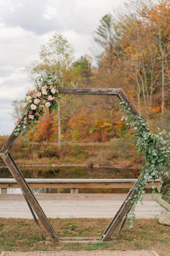 A wooden arbor in front of orange fall foliage. It has a large greenery piece on the right and a smaller floral piece on the left. The flower colors are pink and cream. The florals were created by One Last Avocado Floral Design at Silver Fox Lavender Farm near Wintergreen.