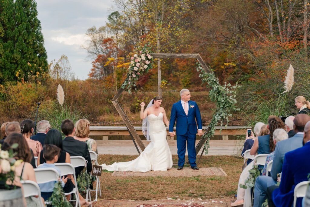 A bride and groom celebrate in front of a wooden arbor with florals on it. The florals were designed by One Last Avocado Floral Design at Silver Fox Lavender Farm in Nellysford, VA.
