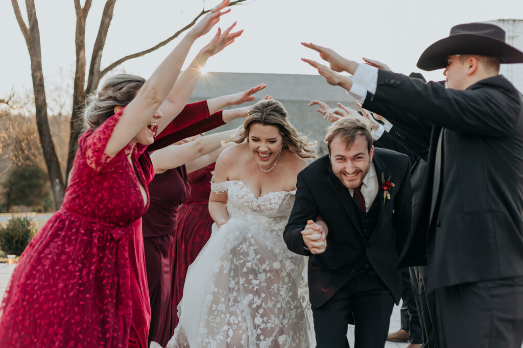 A bride and groom smile happily as they run through a line of their bridesmaids and groomsmen. The groomsmen wear black cowboy hats and the bridesmaids wear varying red dresses during a Leesburg wedding. Wedding venue: 48 Fields Farm. Florist: One Last Avocado Floral Design