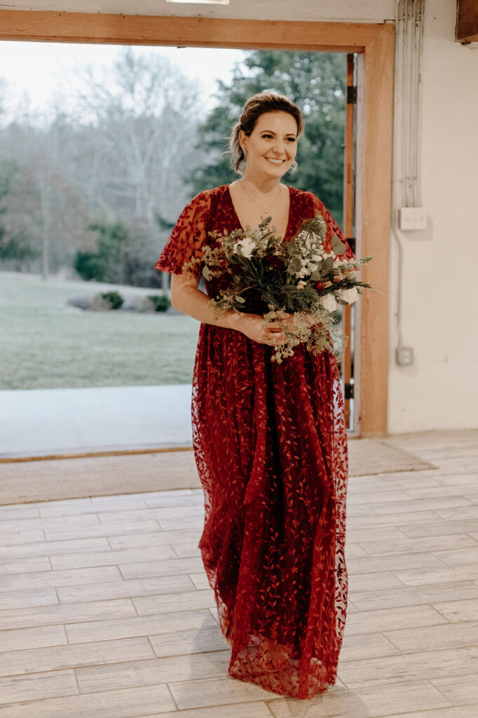 A bridesmaid in a red dress carries a red, white, and green bridesmaid bouquets in a Leesburg wedding. Wedding venue: 48 Fields Farm. Florist: One Last Avocado Floral Design