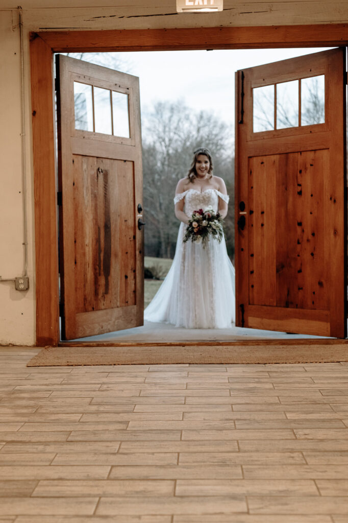 A bride waits to enter her wedding behind large barn doors. She holds a red, white and green Christmas wedding bouquet. Wedding venue: 48 Fields Farm. Florist: One Last Avocado Floral Design