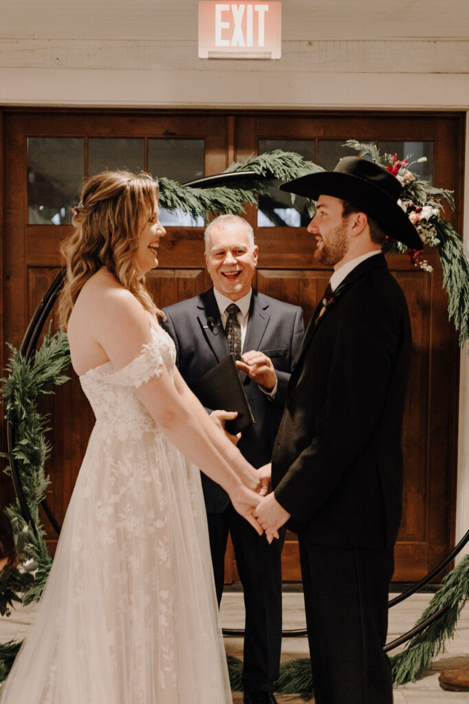 A bride, groom, and officiant stand in front of a wedding arbor with a greenery garland and red and white flowers. Wedding venue: 48 Fields Farm. Florist: One Last Avocado Floral Design