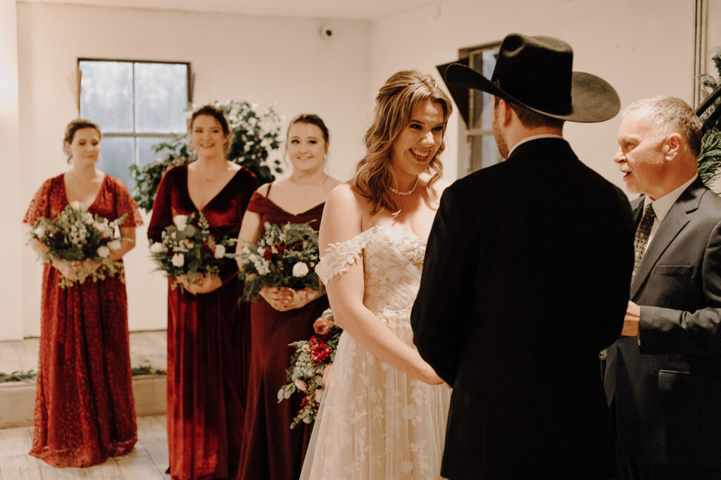 A bride beams at her husband during a Leesburg wedding. Her bridesmaids stand behind her wearing red dresses. Wedding venue: 48 Fields Farm. Florist: One Last Avocado Floral Design