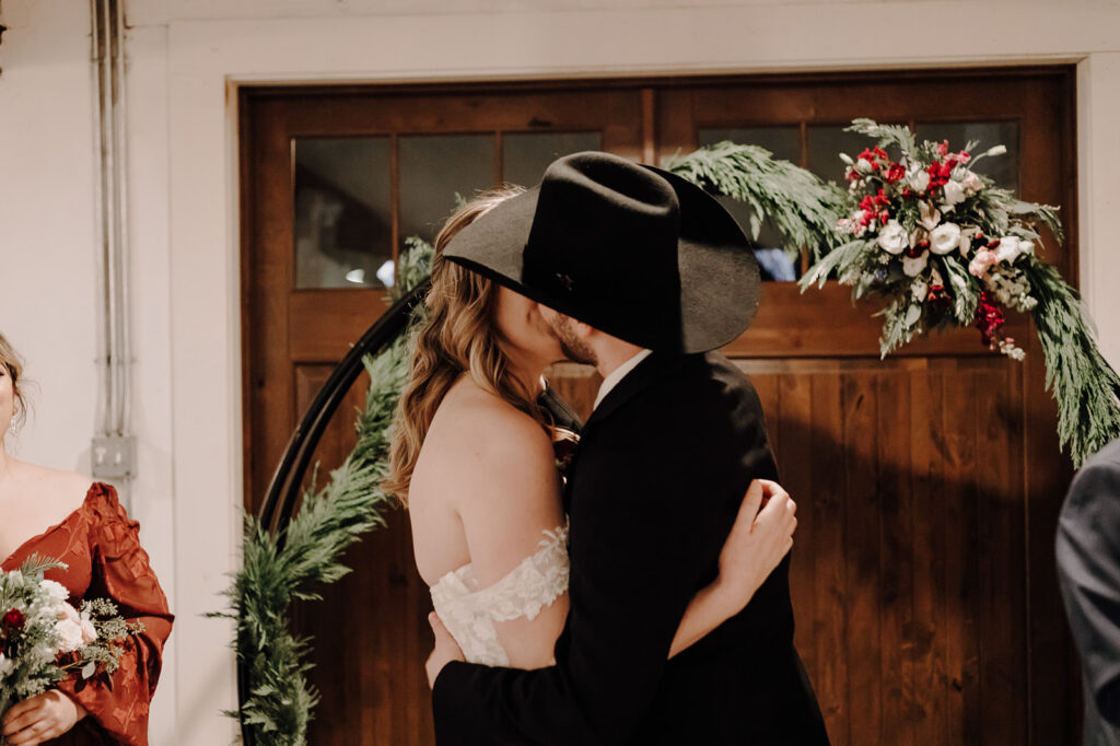 A bride and groom kiss in front of a wedding arbor with a greenery garland and red and white flowers. Wedding venue: 48 Fields Farm. Florist: One Last Avocado Floral Design